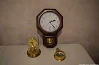 Octagonal dark wood wall clock with pendulum, anniversary clock under glass dome, and a brass hygrometer arranged on a surface.