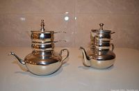 Two silver-plated teapots each with stacked containers above the main pot for storing milk and sugar, showing reflective polished finish and handles.