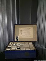 Image of the open blue box showing multiple compartments with metal letters, and a paper sheet with pricing and details attached to the box lid.