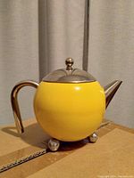 Yellow spherical teapot with stainless steel lid, handle, spout, and three ball feet on a cardboard surface with curtain background.