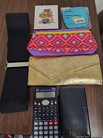 All items in the lot arranged on a wooden surface: two purses (gold metallic and patterned fabric), wide black elastic belt, scientific calculator, framed dried flower decoration, and a blue packaged item.