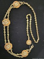 Full view of the linked chain belt showing the five jeweled medallions spaced along the gold-tone double chain on black background.