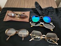 Top view of five pairs of glasses including sunglasses and eyeglasses, displayed on a black surface with two glasses cases.