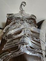 Stack of clear plastic dress hangers with metal hooks photographed on a wooden surface and light-colored couch.