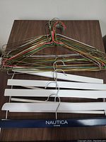 Full view of assorted hangers lying flat on wooden surface showing metal wire hangers in multiple colors and white wooden hangers, including a branded Nautica Home wooden hanger with clips.