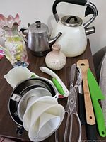 Overview of all kitchen items on table including teapots, ceramic vases, strainers, peelers, spatulas, and clip.