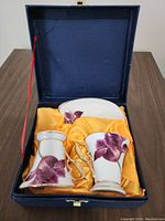 Cup and saucer with purple floral design inside blue presentation box with yellow satin lining.