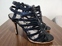 Side front view of black strappy high heel sandal shoe showing multiple straps with faux snakeskin pattern on wooden surface.