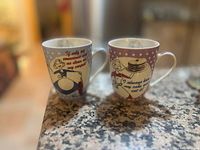 Two decorated ceramic teacups placed on a granite countertop with humorous vintage-style prints visible on the sides.