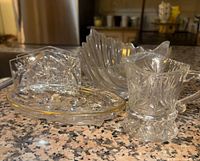 Clear crystal glass serving plates of various shapes and a small matching pitcher with handle, placed on a granite countertop.