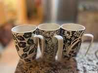 Photo of three tall ceramic teacups with black and white floral and leaf designs on a countertop.
