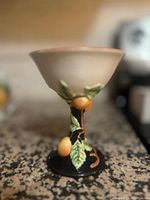 Photo of a ceramic chalice with a wide rim and a stem decorated with green leaves and orange fruit details, placed on a speckled countertop.