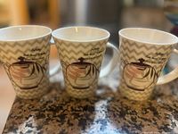 Three ceramic teacups with zigzag pattern and coffee pot graphic on counter.