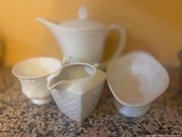 Photo of white ceramic tea set items on countertop including teapot, creamer, sugar bowl, and pedestal dish