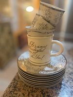 Stack of 6 white plates with silver snowflake designs topped by two white mugs, one with festive winter text.