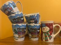 Photo of six colorful ceramic mugs with winter and Christmas themed prints including a snowman and moose, arranged in a pyramid style.