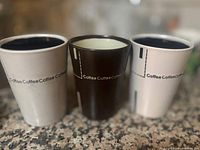 Three coffee mugs arranged on a granite countertop: two white mugs on the sides and a black mug in the center. Each mug has 'Coffee' repeated in text near the top.