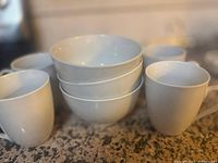 Photo showing four white coffee cups and four white bowls stacked on a countertop.