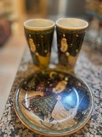 Photo of one antique Christmas decorative plate and two tall matching Christmas-themed glass teacups with detailed holiday illustrations on dark backgrounds with gold accents.