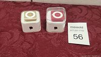 Two Apple iPod Shuffles in plastic cases side by side on a red textured fabric surface with a lot number tag nearby