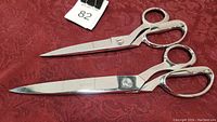 Two pairs of shiny Gingher scissors on a red fabric background, showing full length and details.