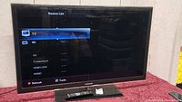 Samsung 46 inch TV powered on displaying source list menu, TV sitting on table with remote in front.