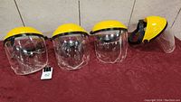 Four yellow hard hat face shields with clear visors lined up on a red cloth surface.