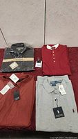 Four men's shirts displayed, two folded and two laid out flat on a red surface against a tiled wall. All shirts have new tags.