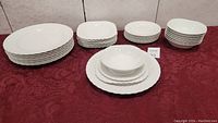 Photo of all 32 pieces of Johnson Brothers Snow White Regency dinnerware arranged in stacks by type on red tablecloth.