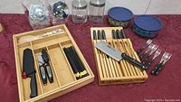 Photo showing two bamboo utensil trays with black-handled knives, cutters, forks, spoons, and chopsticks arranged inside compartments, plus two clear canisters with silver lids in background.