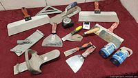 Photo shows full array of drywall tools on red cloth background including taping knives, putty knives, measuring tape, drywall square, and joint compound containers.