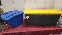 Photo showing both blue storage bin and large black tote with yellow lid side by side on table.