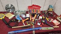 Overview photo showing the full assortment of tools and hardware on a red surface including pliers, screwdrivers, hacksaw, measuring tape, wire brush, wall clock, and fly swatter.