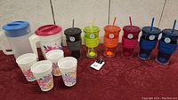 Photo showing colorful travel cups with silicone grips, lids, and straws, along with two white plastic juice pitchers and four small disposable floral cups.