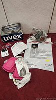 Uvex safety goggles placed on their box alongside 3M respirator, instruction sheet, and two pink filters in open packaging on a red surface.