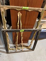 Two antique wooden window frames leaning against a wooden panel; cream colored ornate frame with two matching sashes and plain black window frame with some paint wear and chipping.