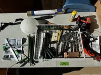 Top-down view of tools on a table including a glass bottle cutter with adapters, cordless drill, clamp, green clamps/stakes, glue tube, and a white glass lampshade.