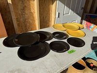 Nine plates arranged on a table in sunlight, five black plates in varying sizes and four yellow plates of similar sizes. Plates are round with a grooved pattern on the rim, matte black and glossy yellow finishes.
