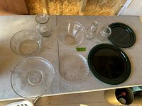 Top down photo showing assortment of clear glass bowls, plates, and small bottles, plus dark green glass plates, on a light-colored table.