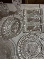 Various clear glass tableware items including trays, plates, bowl, candy dish, decanter, and vase on white surface