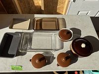 Top down photo showing all dishes laid out on table in sunlight, includes clear Pyrex glass dishes and multiple brown stoneware casseroles and baking dishes with lids.