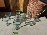Six margarita glasses, a clear glass pitcher with green rim, and a multicolored woven basket shown on a table from side angle.