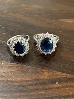 Two blue stone rings side by side on wood surface. Both rings feature large oval dark blue faceted stones surrounded by clear round accent stones in a halo design.