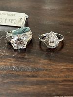 Two sterling silver rings side by side on wood surface, one with large round clear stone, the other with pear-shaped clear stone with surrounding smaller stones