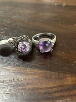 Two sterling silver rings with purple stones on a wooden surface, one with a round faceted stone in a decorative setting, the other with a cushion-cut stone and textured band.