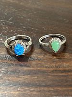 Two sterling silver rings side by side on a wooden surface. One ring has an oval vibrant blue stone, the other a pear-shaped green stone with accent stones.