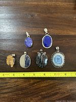 Top-down view of six silver pendants arranged on wooden surface next to a measuring tape for size reference.
