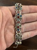 Close-up photo of silver bracelet held in hand, showing linked gemstone-studded segments in multiple colors.
