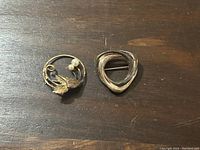 Two sterling silver brooches on wood surface. Left brooch is circular with leaf and pearls, right brooch is twisted triangular shape.