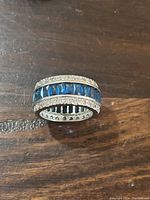 Close-up photo of the sterling silver infinity band ring on a wood surface showing blue square-cut stones in the center and small white stones bordering them.